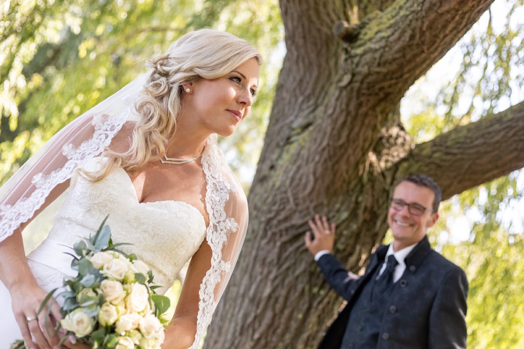 Braut mit Brautstrauß, im Hintergrund der Bräutigam an einem Baum